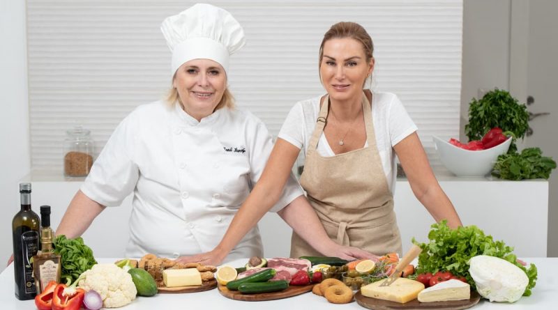 Two female chefs with variety of fresh ingredients in a kitchen setting, showcasing culinary preparation.